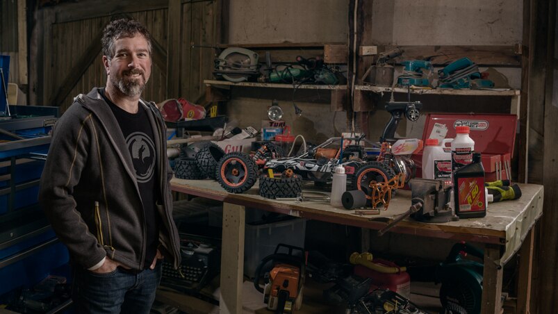 A man stands in a wooden shed next to a workbench, on which sits remote control cars.