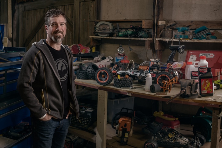 A man stands in a wooden shed next to a workbench, on which sits remote control cars.