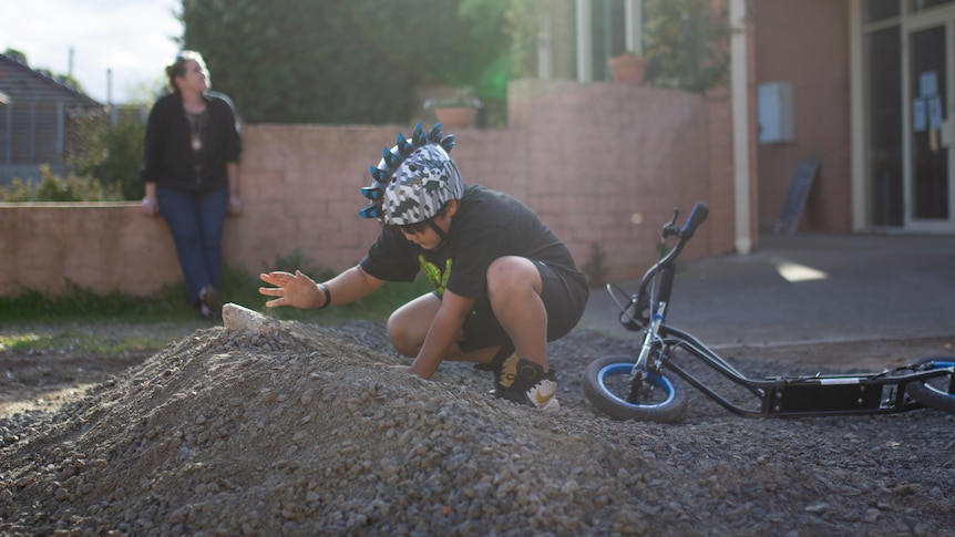 A boy wearing a bike helmet makes a jump for his scooter while his mum watches in the background.