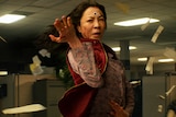 Papers fly in office as Chinese-American woman with long dark hair wearing floral shirt and red vest performs kung fu move.