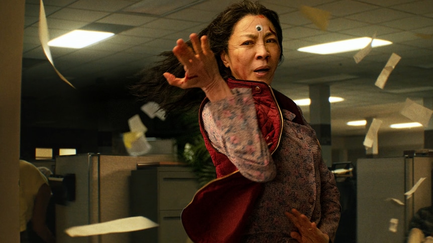 Papers fly in office as Chinese-American woman with long dark hair wearing floral shirt and red vest performs kung fu move.