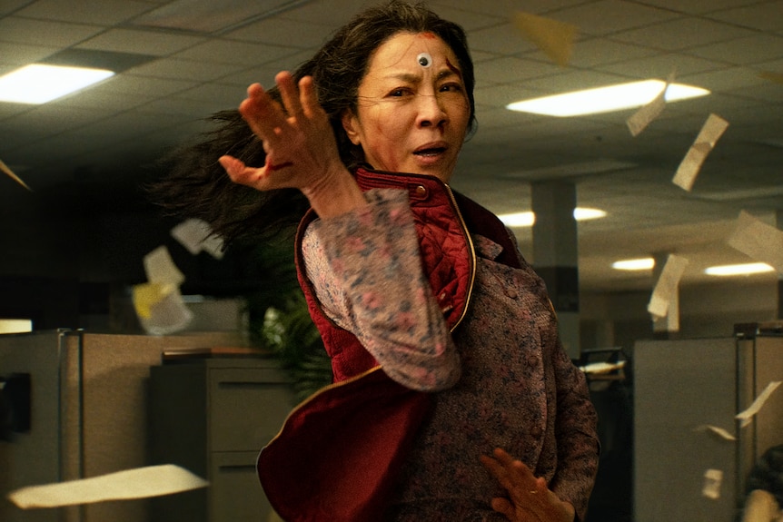 Papers fly in office as Chinese-American woman with long dark hair wearing floral shirt and red vest performs kung fu move.