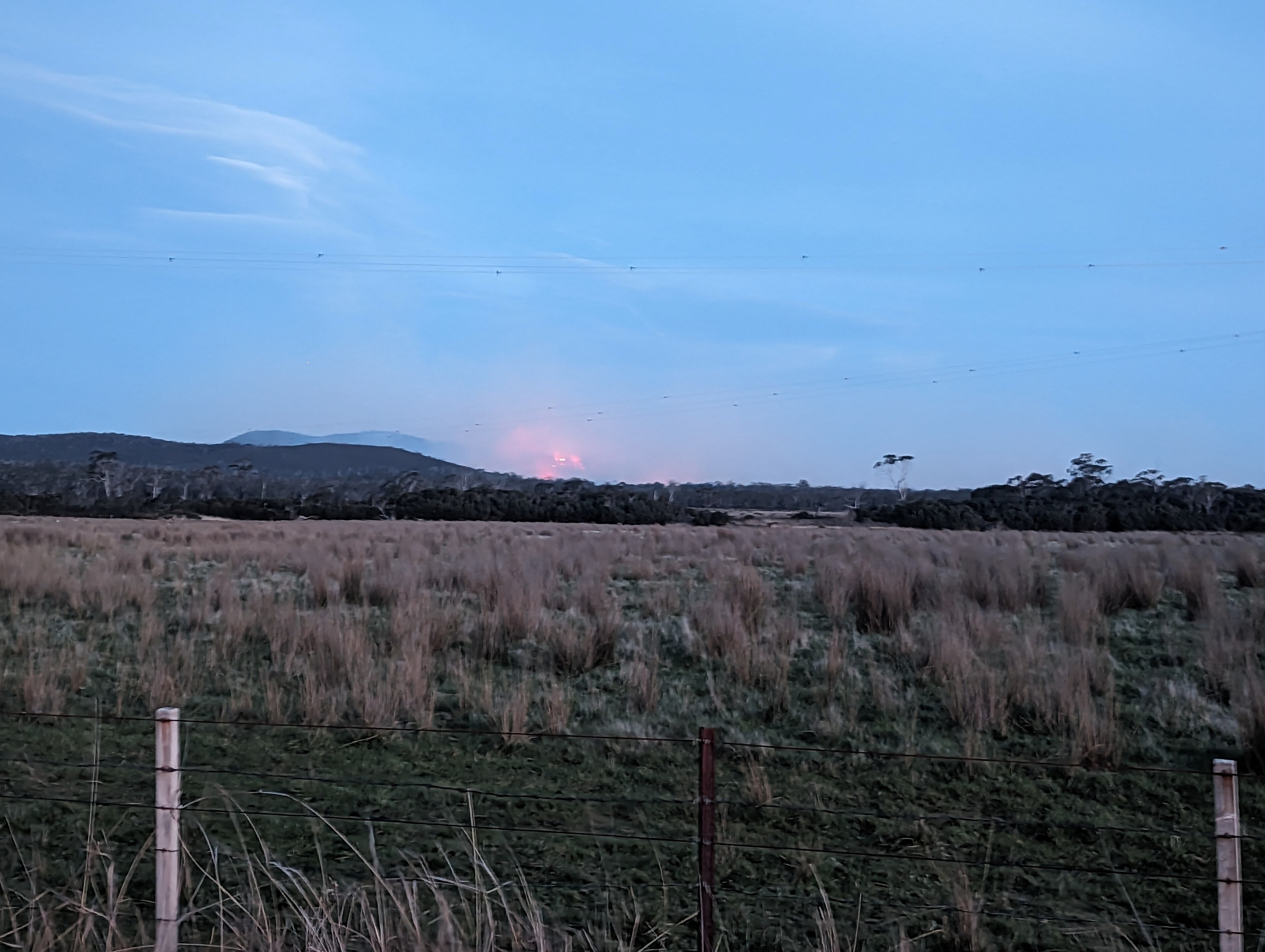 A red glow and smoke rise above a hill at dusk with paddocks in the foreground.