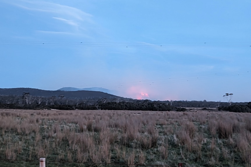 A red glow and smoke rise above a hill at dusk with paddocks in the foreground.