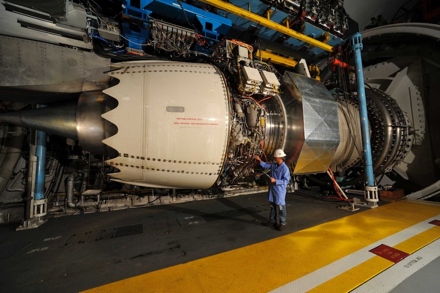 In a large military facility, a large jet engine with frill-like ends is exposed and plugged into a litany of different systems.