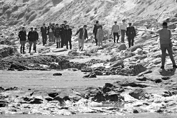 A large group of people walking along a rocky beach with sand dunes in the background.