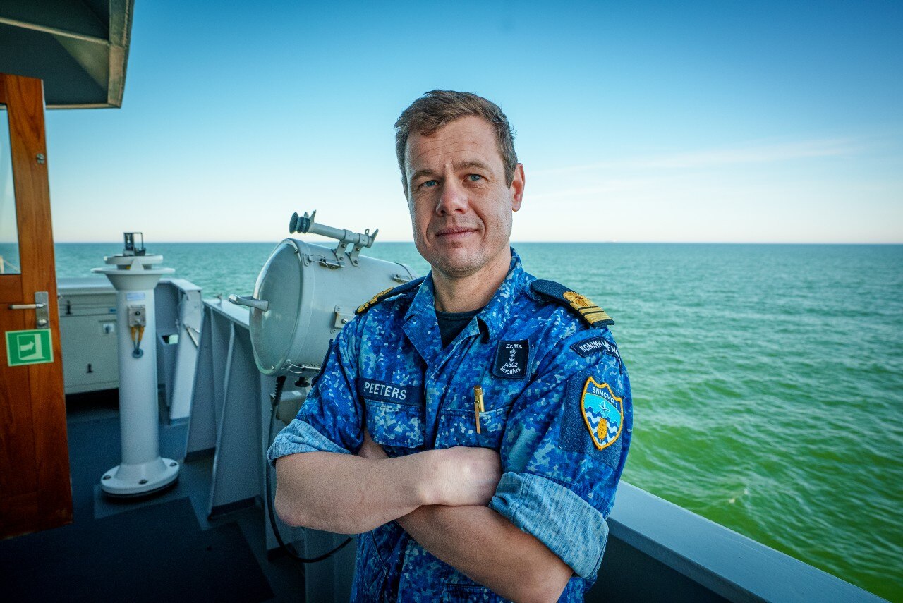 A man in a navy uniform with his arms folded standing on the deck of a ship, looking on.