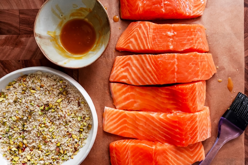 Six raw salmon fillets on a chopping board beside a bowl of pistachio bread crumbs and honey marindade.