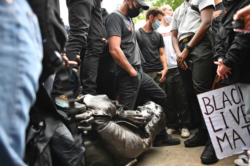 A statue of a man lies on the ground surrounded by protesters
