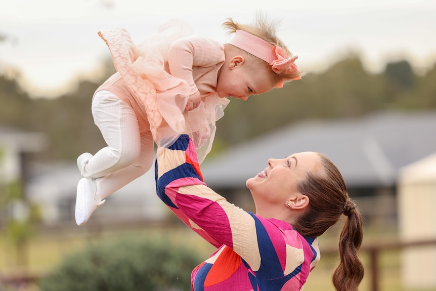a mother holds her baby daughter high in the air, they are both looking at each other and smiling