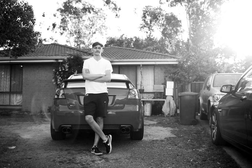 A tall young man stands with arms folded leaning against a car in front of his suburban home in western Sydney.