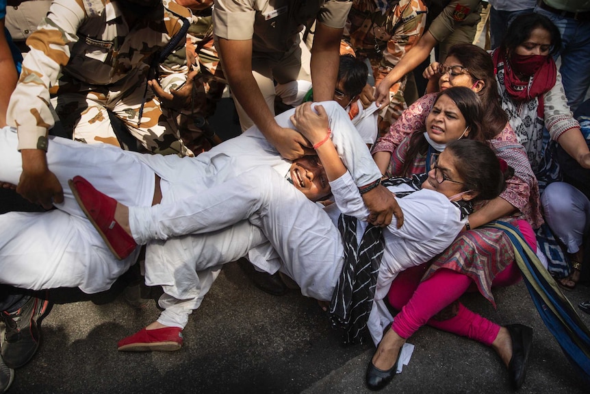 A group of people, including a number of women, hold each other in a group on the ground as soldiers attempt to detain them