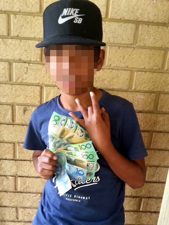 A young boy wearing an oversized nike hat holds up a fan of $100 and $10 notes while pulling a 'west side' sign with his fingers