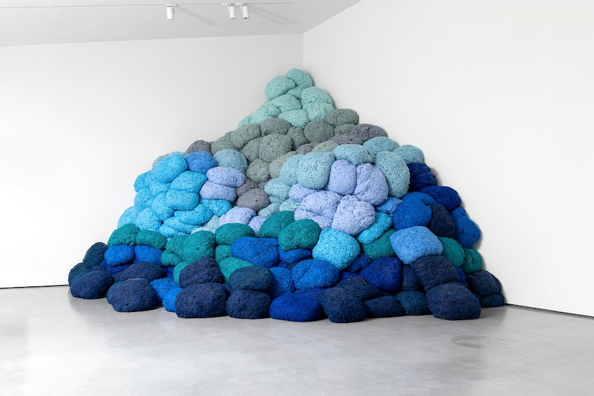 A pile of blue fabric bundles, in different shades from royal blue to pale blue, piled in the corner of a white gallery.