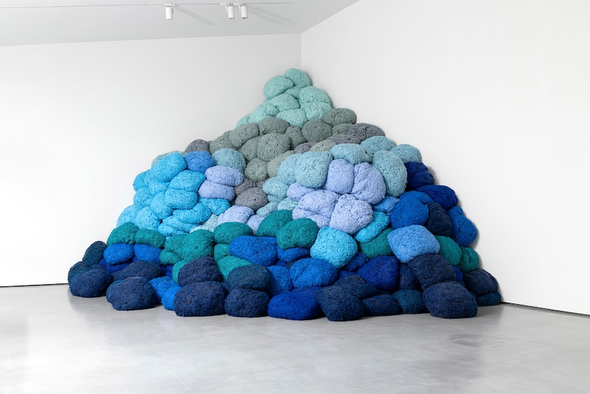 A pile of blue fabric bundles, in different shades from royal blue to pale blue, piled in the corner of a white gallery.