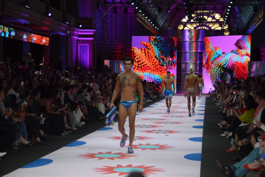 Men wearing swimwear featuring bright blue and white are walking up and down a white runaway.