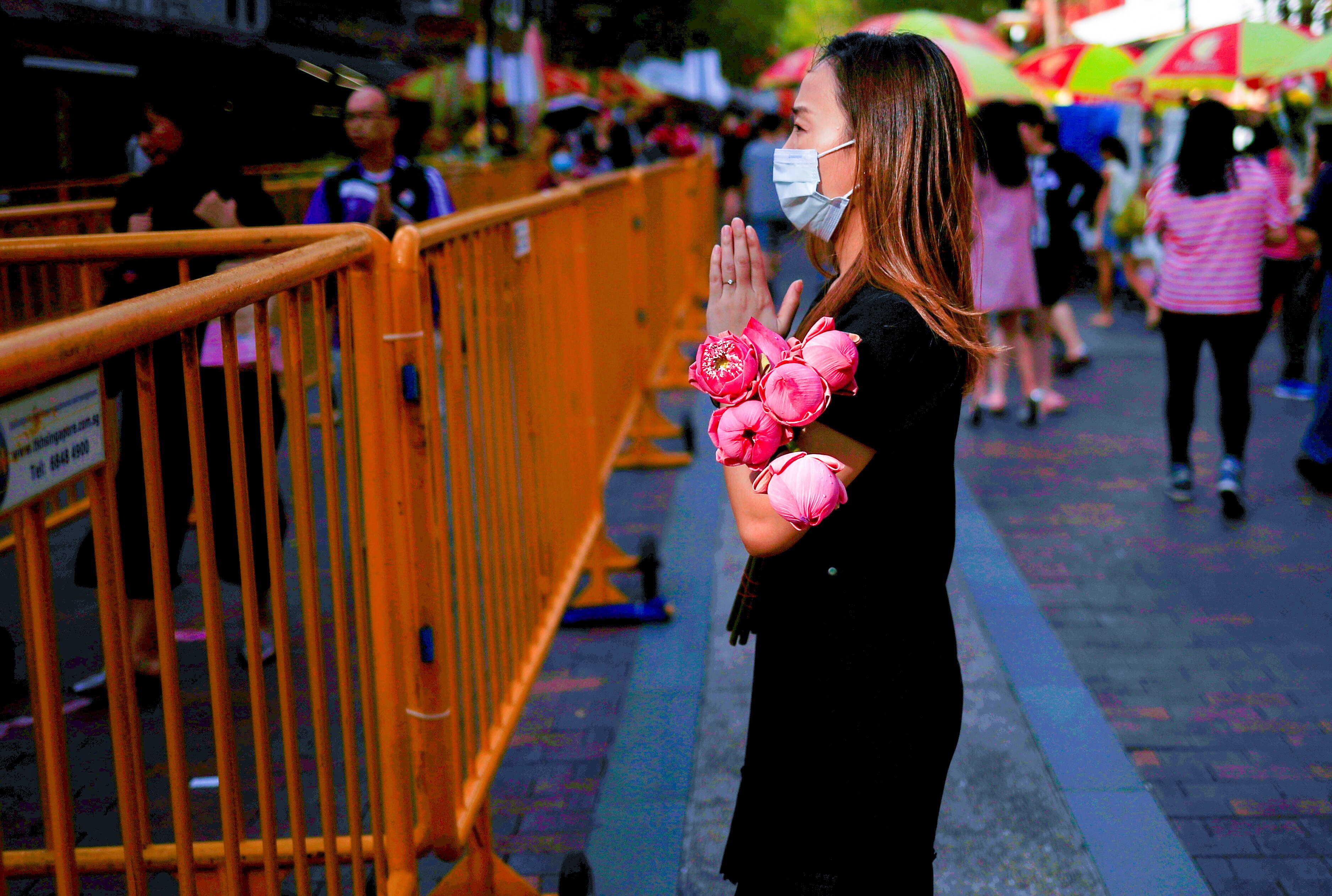A woman in a mask holds her hands in prayer with a bunch of flowers in the crook of her arm