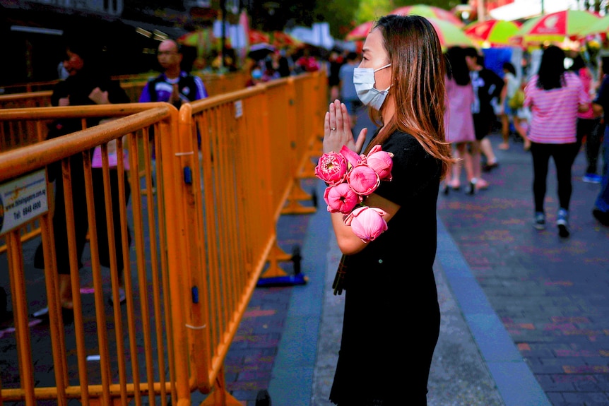 A woman in a mask holds her hands in prayer with a bunch of flowers in the crook of her arm