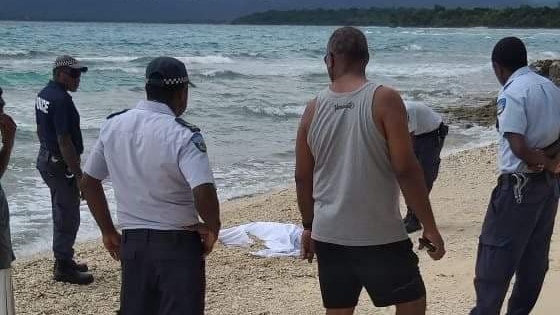 Several men and officials standing around a body draped with white cloth on a beach.
