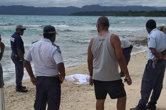 Several men and officials standing around a body draped with white cloth on a beach.