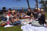 A group of people on a picnic rug at a beach with drinks in hand.