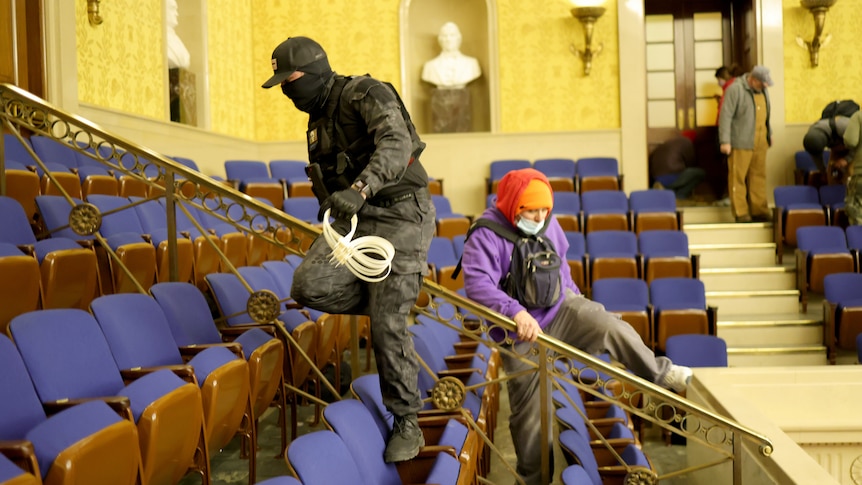 A masked man in military gear holds zip-tie handcuffs in the senate chamber of the US Capitol