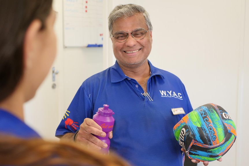 A man with a blue WYAC shirt holds a ball and a water bottle while smiling