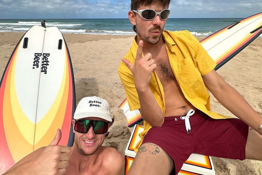 Two men wearing sunglasses on a beach with "better beer" written on a surfboard