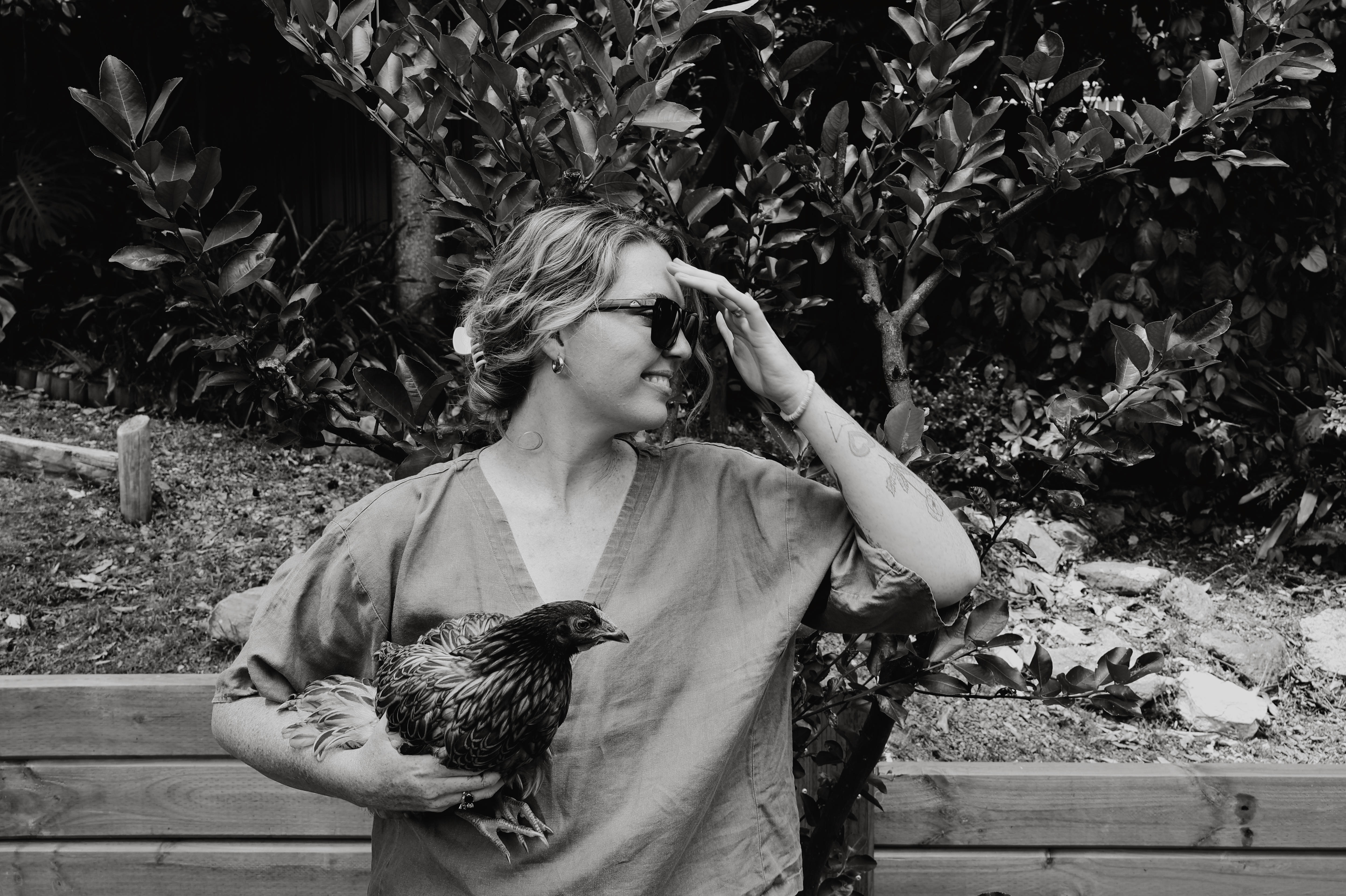 Black and white photo of Amy Thunig looking sideways and smiling with a chook under her arm. 