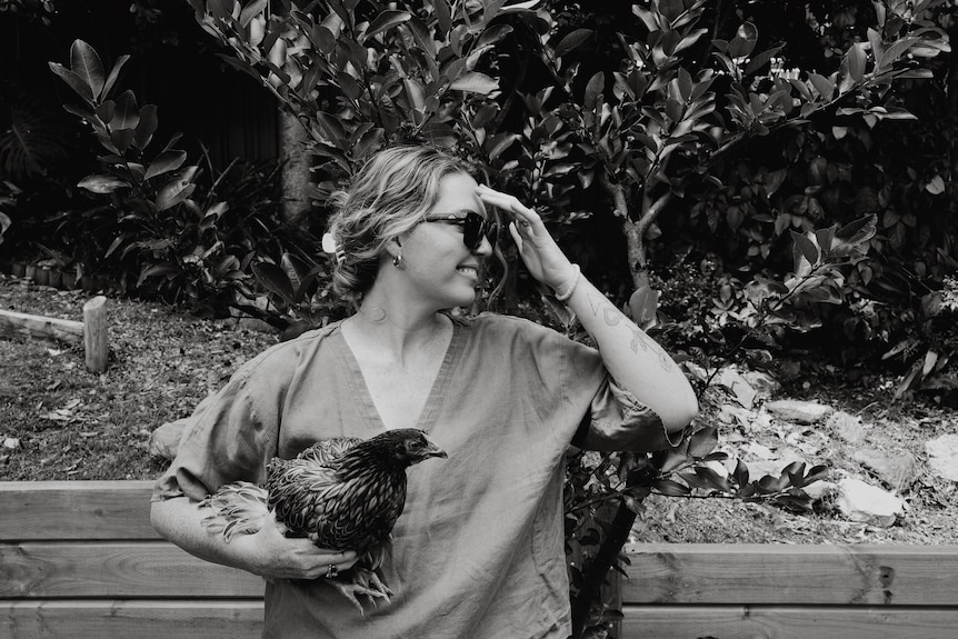 Black and white photo of Amy Thunig looking sideways and smiling with a chook under her arm. 