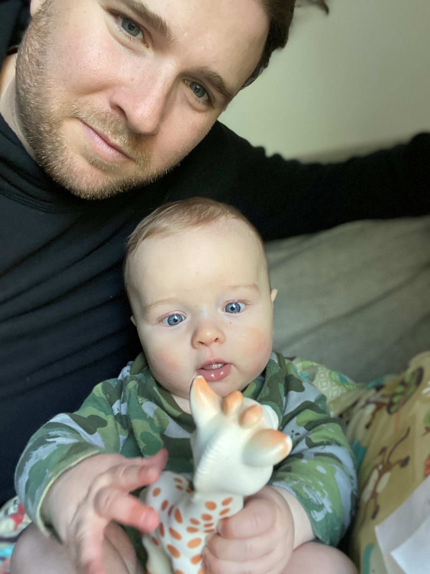 A smiling man holds his arm around an infant who is happily squeezing a toy giraffe.