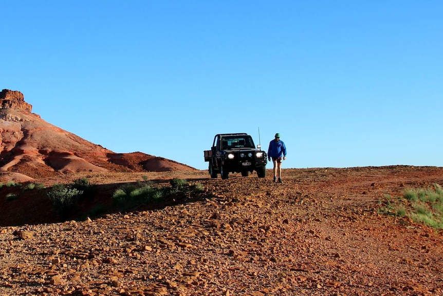 Jamie Black walks from his car down a gravel road. In the background, a big red sand jump up.