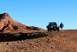 Jamie Black walks from his car down a gravel road. In the background, a big red sand jump up.