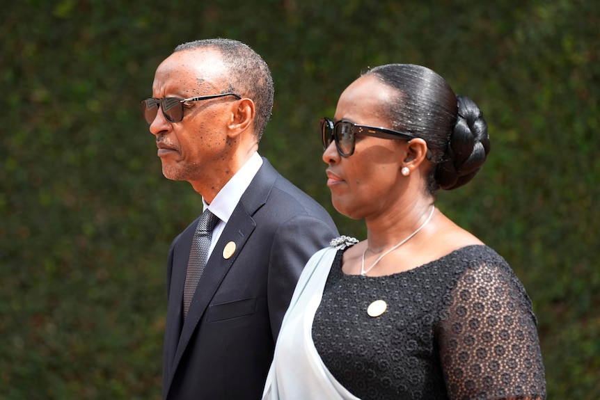 An older African man in a suit walks next to an African woman in a dress, both wearing sunglasses.
