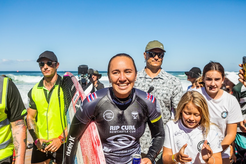 A young female surfer smiles as people stand around her at the beach.