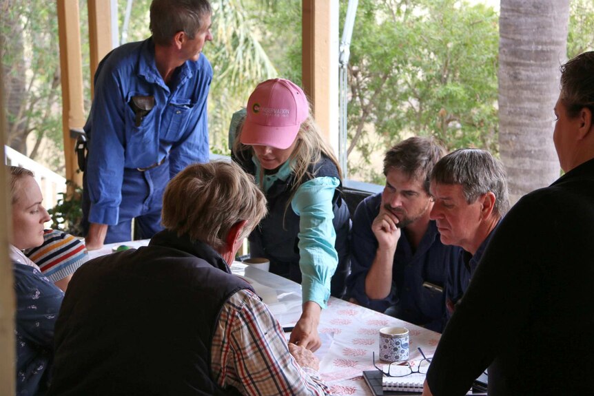 Seven Brigalow locals meet at Terry Dalgliesh's homestead and look at documents on a table on the verandah.