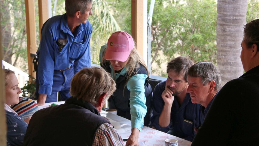 Seven Brigalow locals meet at Terry Dalgliesh's homestead and look at documents on a table on the verandah.