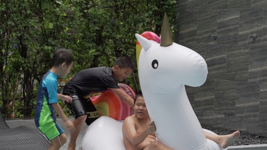 Three children play on a blow up unicorn toy while jumping in the pool.