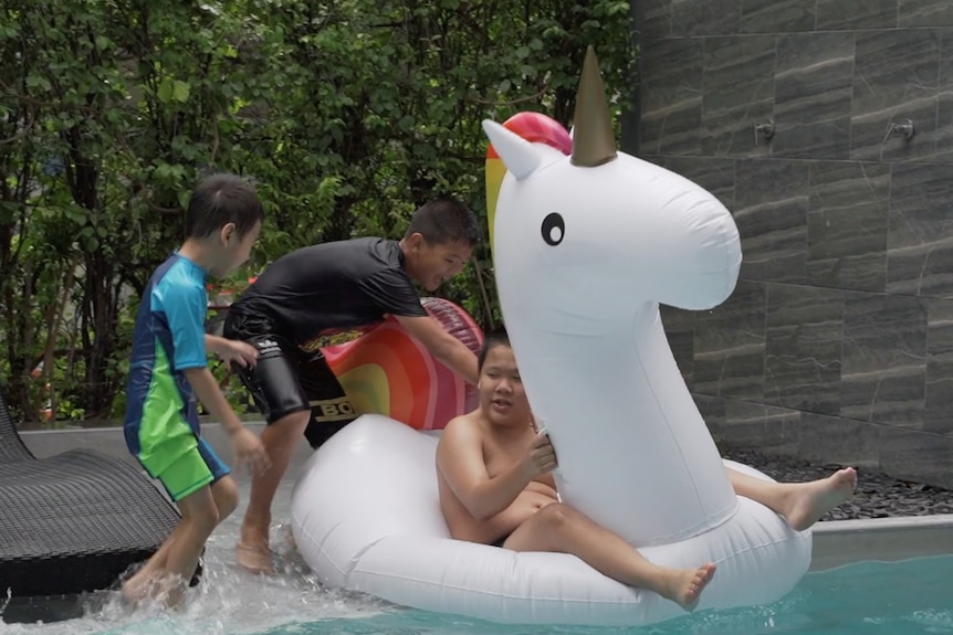 Three children play on a blow up unicorn toy while jumping in the pool.