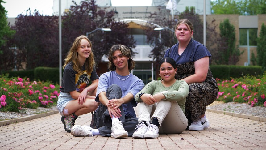 Four young people sitting on a paved footpath in front of a building.