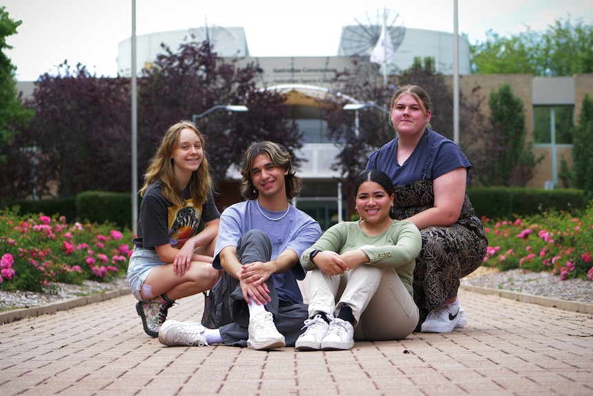 Four young people sitting on a paved footpath in front of a building.