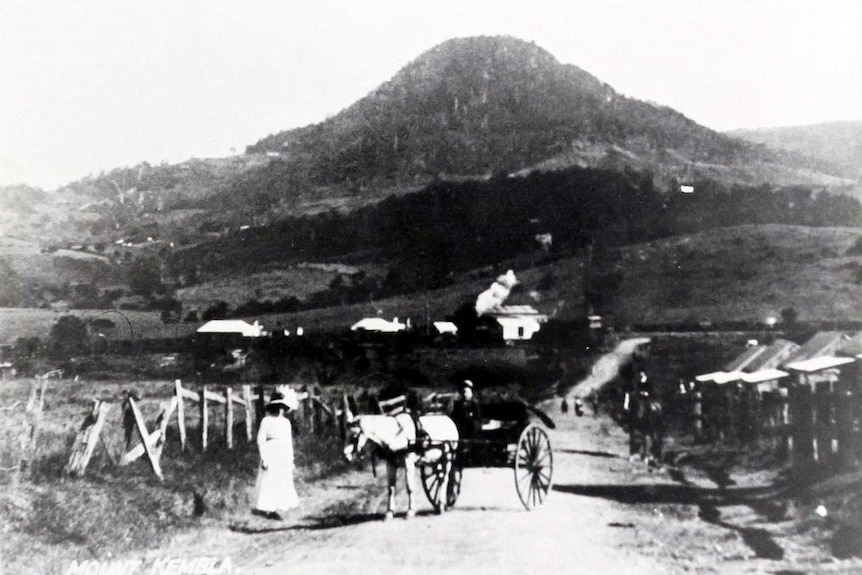 Black and white image of a woman standing next to a horse and cart on a dirt road with Mount Kembla in the background.