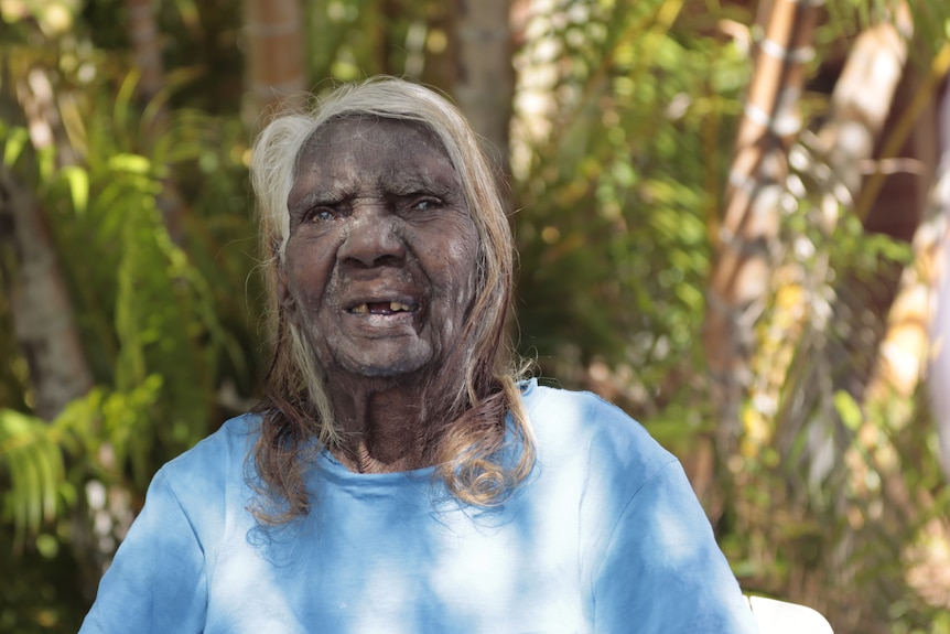 a woman with long grey hair and missing teeth is smiling to the camera. 