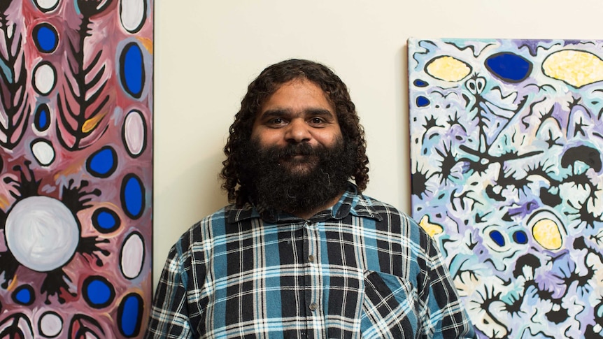 Indigenous artist Cedric Varcoe stands in the middle of his two paintings hanging in the Murray Bridge Regional Gallery