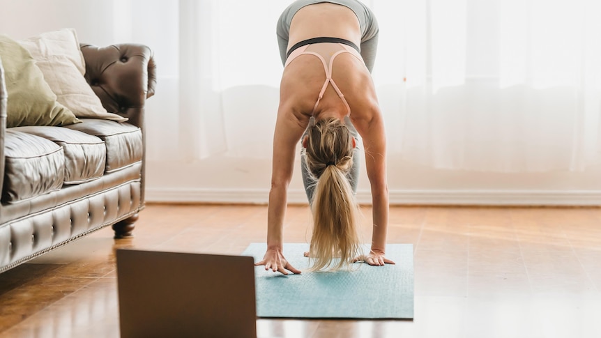 A woman does yoga in front of her computer in her lounge room.