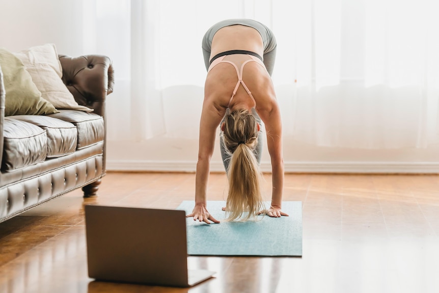 A woman does yoga in front of her computer in her lounge room.