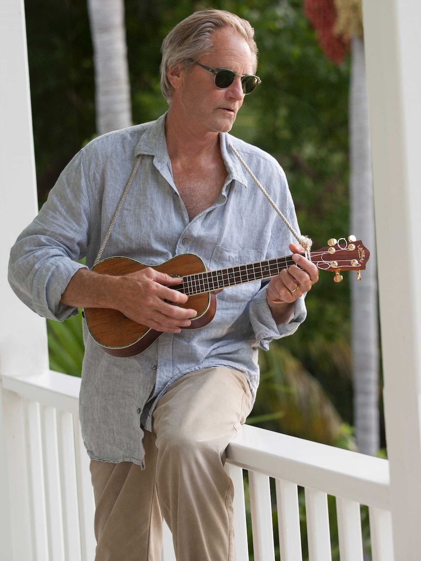 Sam Shepard leans on a railing playing the ukulele.