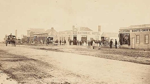 A sepia photo of a town in the 1860s, with horses and carts and old wooden buildings.