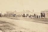 A sepia photo of a town in the 1860s, with horses and carts and old wooden buildings.
