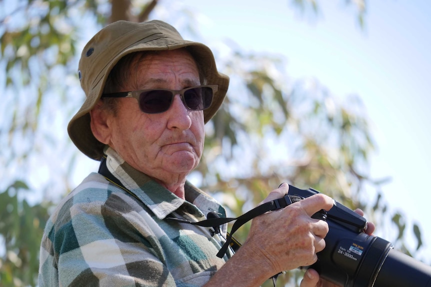 A man in sunglasses and a fishing hat looks at the camera while holding a camera with a very long lens.